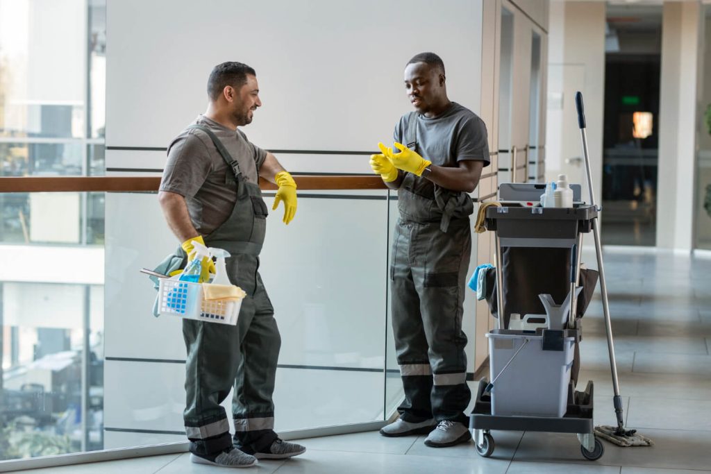 A team of professional cleaners in gray uniforms tidying up a modern home. One cleaner is vacuuming the living room, another is dusting shelves, and another is sanitizing the kitchen counter, creating a fresh and welcoming space.